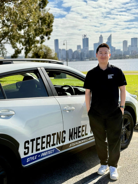 Person standing next to a car with 'Steering Wheel Wraps Perth' branding, Perth city skyline in the background